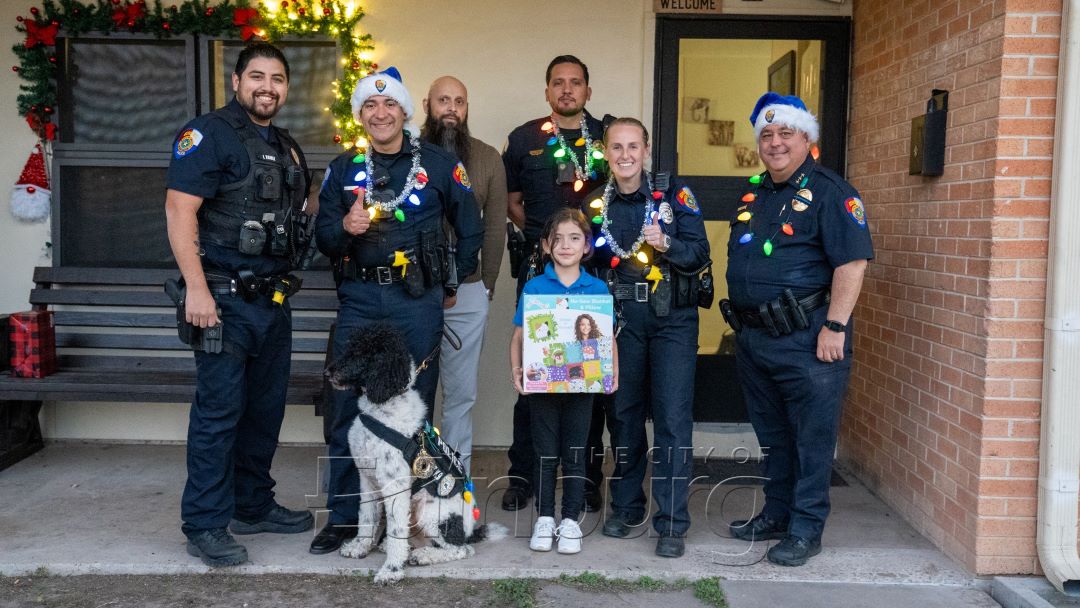 Group of uniformed police officers standing with children and a dog outside a residence, with holiday decorations visible near the entrance. The children are holding items and posing for a photo.