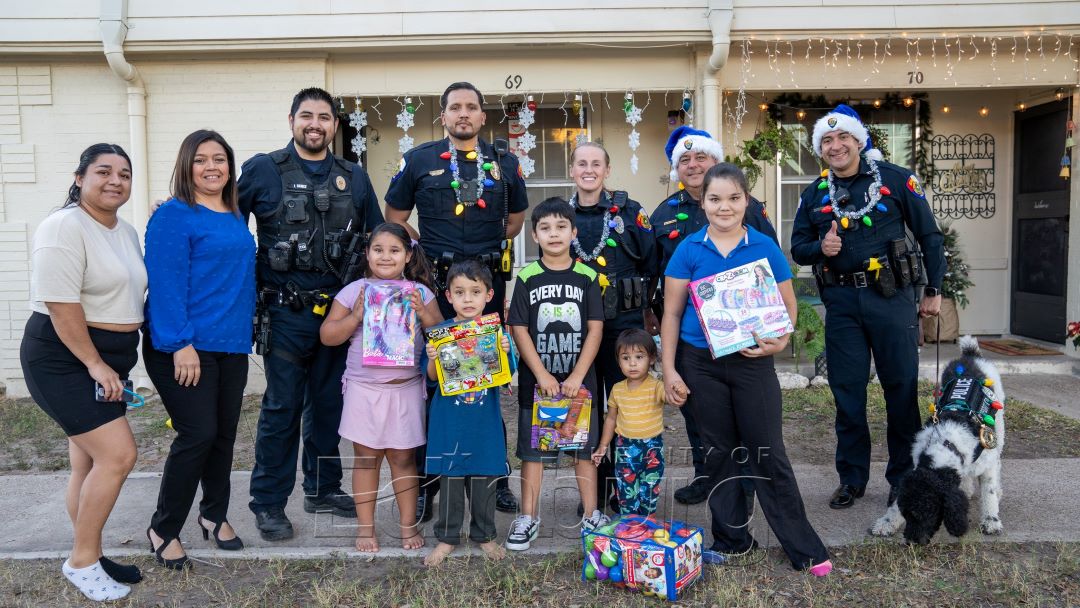 Police officers standing with several children and adults outside a home, with children holding wrapped gifts and smiling for a group photo.