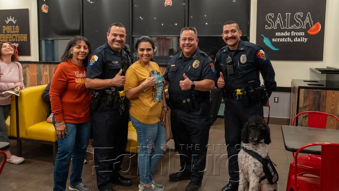 Three smiling Edinburg police officers and two women give thumbs-up signs while standing inside a restaurant. A black-and-white service dog sits in the foreground next to the group