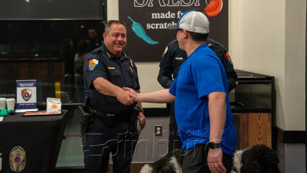 Uniformed police officer shaking hands with a man wearing a cap inside a business establishment, with a table and signage visible in the background.