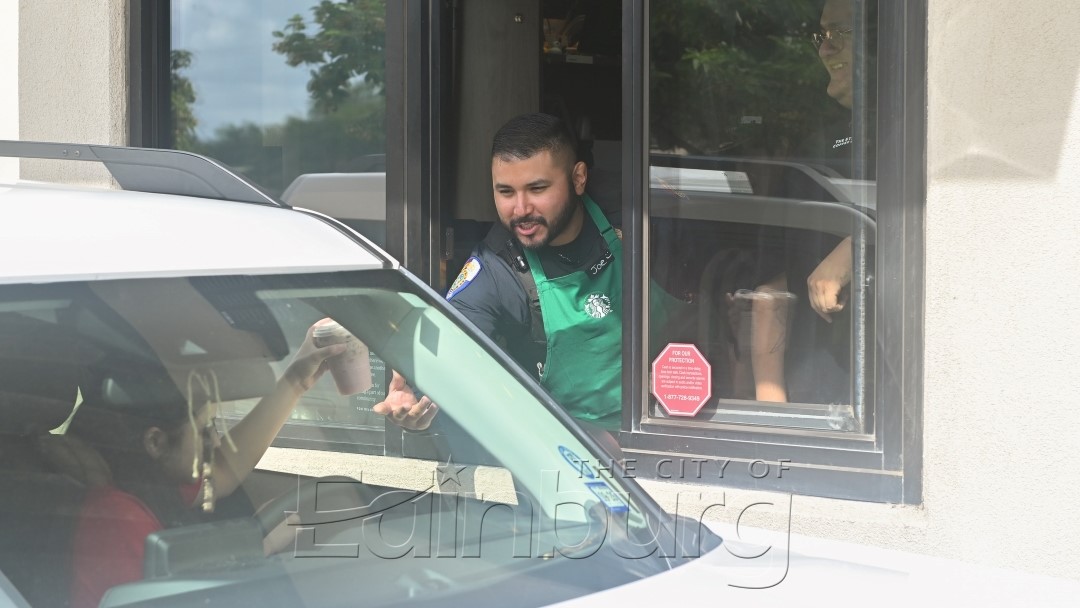 Police officer working at a drive-through window handing an item to a person inside a vehicle, with the car partially visible outside.