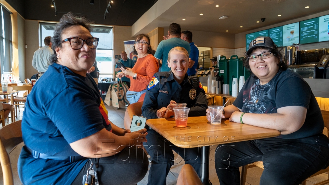 Police officer seated at a table inside a coffee shop with two community members, all smiling, with drinks and caf&eacute; seating visible in the background.