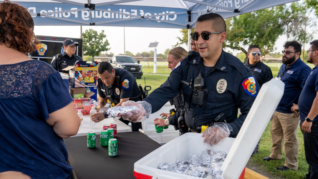 Police officer wearing gloves handing food to a community member across a table, with other officers and supplies visible at an outdoor event.