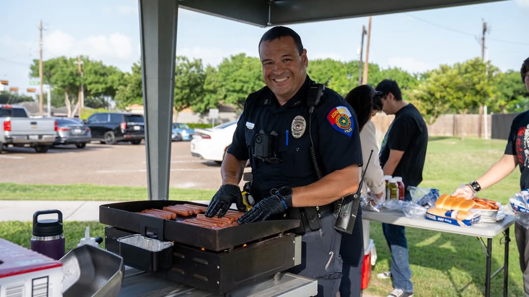 Police officer smiling while cooking food on a grill outdoors, with tables, supplies, and other people visible in the background.
