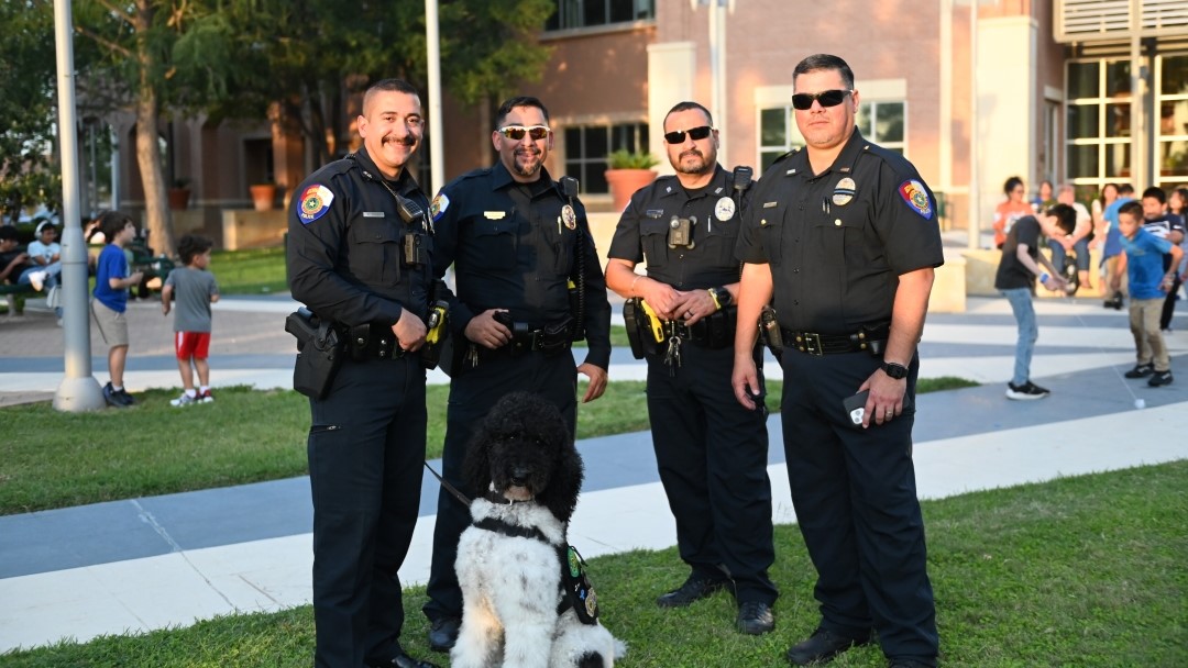 Four Edinburg police officers stand together on a paved walkway in a park-like setting during the day, with a black-and-white service dog sitting in front of them. In the background, several children are playing on the grass and near park benches.